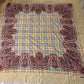 A large square scarf laid flat on a wood floor, featuring a beige plaid center with an ornate red, black, and white paisley border and a solid beige bottom border.