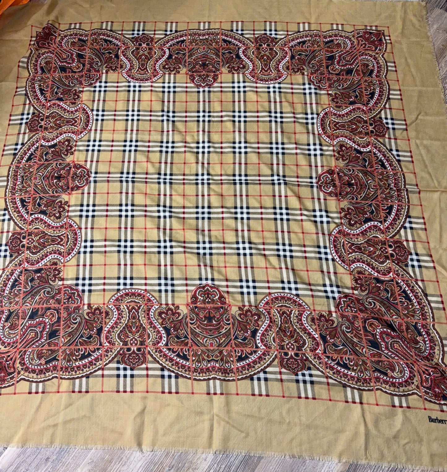 A large square scarf laid flat on a wood floor, featuring a beige plaid center with an ornate red, black, and white paisley border and a solid beige bottom border.