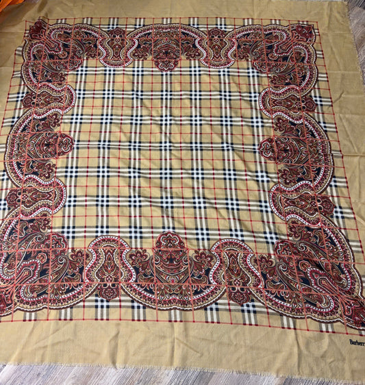 A large square scarf laid flat on a wood floor, featuring a beige plaid center with an ornate red, black, and white paisley border and a solid beige bottom border.